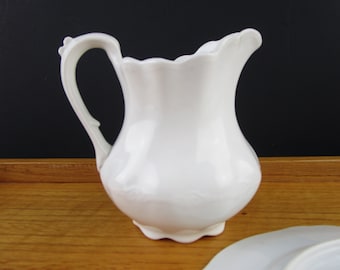May include: A white ceramic pitcher with a curved handle and a scalloped rim. The pitcher has a bulbous body and sits on a small foot. A white plate is partially visible in the foreground. The pitcher is likely used for serving beverages.