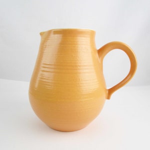 May include: A yellow ceramic pitcher with a rounded body and a handle. The pitcher has a smooth, slightly textured surface.