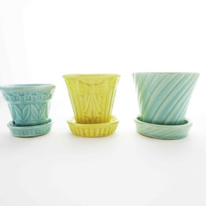 May include: Three ceramic flower pots in shades of blue and yellow, each with a matching saucer. The pots have different designs: one with a leaf pattern, one with a bow, and one with a spiral texture. They are set against a white background.