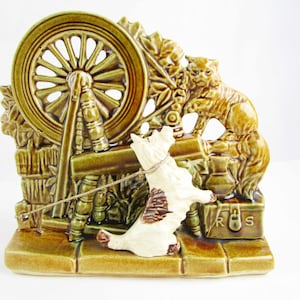 May include: A ceramic planter shaped like a spinning wheel with a cat and a dog figurine. The planter is glazed in a green and brown color scheme. The dog is white with brown spots and is pulling on a string attached to the spinning wheel. The cat is brown and is sitting on a box with the letters "R.S." on it.