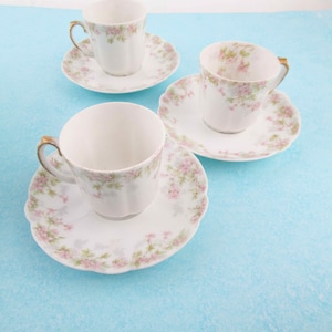May include: Three white teacups and saucers with a floral pattern of pink flowers and green leaves. The teacups have gold-colored handles. The saucers have a scalloped edge. The teacups and saucers are on a light blue surface.