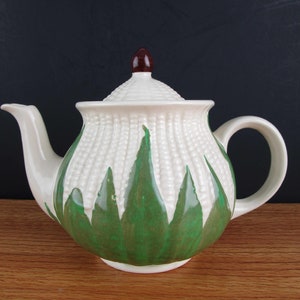 May include: White ceramic teapot with a green and white corn design. The teapot has a brown knob on the lid.