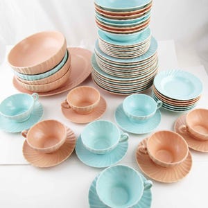 May include: A collection of vintage pastel-colored dishware, including bowls, plates, and teacups with saucers. The set features a mix of light blue and peach-colored pieces, arranged on a white surface. The dishes have a ridged design.