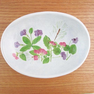 May include: White oval ceramic dish with a pressed flower design. The design features pink and purple flowers, green leaves, and a pine needle.