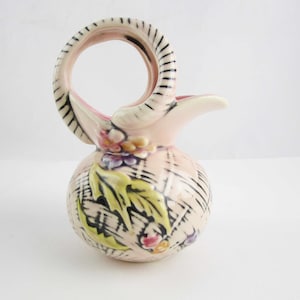 May include: A small pink ceramic pitcher with a black and white patterned handle. The pitcher has a floral design with green leaves and pink, yellow, and purple flowers.
