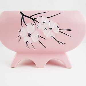May include: A pink ceramic planter with a floral design. The planter has three legs and is decorated with white flowers and black stems.