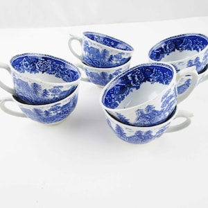 May include: Set of six white ceramic teacups with a blue landscape pattern. Each cup has a curved handle and a matching saucer design. The cups are stacked and arranged on a white surface.