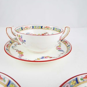 May include: A white porcelain soup bowl with a floral pattern and red, blue, and yellow accents. The bowl has two handles and is sitting on a matching saucer.