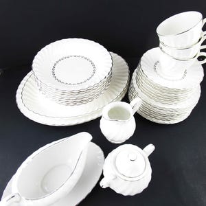 May include: A white porcelain tea set with silver trim. The set includes plates, saucers, cups, a creamer, a gravy boat, and a teapot. The plates and saucers have a scalloped edge and a delicate floral design.