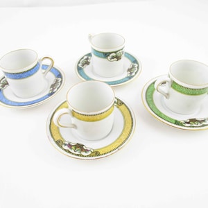May include: Four white espresso cups and saucers with gold trim and colorful bands of blue, green, and yellow. Each saucer features a decorative cloud-like design. The cups have gold handles.
