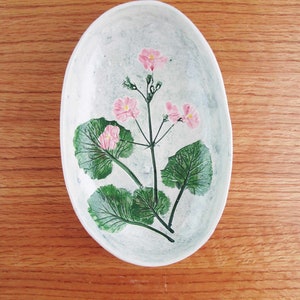 May include: A white oval ceramic dish with a floral design. The dish features a pink flower with green leaves painted on the surface.
