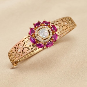 Peut inclure: Un bracelet jonc articulé doré avec un motif en filigrane ajouré. Le centre présente une grappe florale de pierres magenta ovales entourant une pierre centrale transparente, avec le texte BQ0702-RD-75 inscrit.