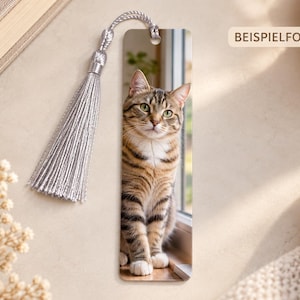 Könnte beinhalten: Ein rechteckiges Lesezeichen mit einem Fotodruck einer braun-schwarzen getigerten Katze, die an einem Fenster sitzt. Das Lesezeichen hat eine silberfarbene Kordelquaste, die oben durch ein kleines Loch befestigt ist. Der Text BEISPIELFOTO ist sichtbar.