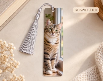 May include: A rectangular bookmark featuring a photographic print of a brown and black tabby cat sitting by a window. The bookmark has a silver-colored cord tassel attached to the top through a small hole. The text BEISPIELFOTO is visible.