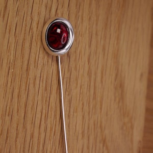 May include: A silver-toned lapel pin with a round, red gemstone center. The gemstone has a swirling pattern. The pin is set against a wooden background.