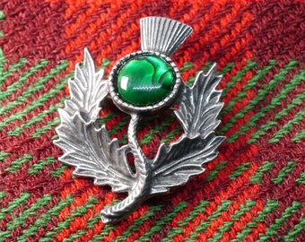 May include: A silver thistle brooch with a green abalone shell center. The brooch is on a red and green plaid fabric.