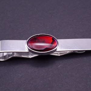 May include: A silver tie clip featuring an oval, deep red gemstone. The gemstone is set in a silver bezel. The clip has a simple, elegant design. The overall aesthetic is one of understated sophistication.