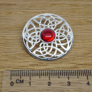 May include: Silver Celtic knot brooch with a red gemstone in the centre. The brooch is approximately 5 cm in diameter.