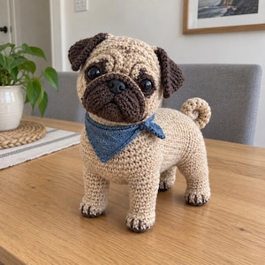 May include: A crocheted pug figurine featuring a tan body, dark brown ears, and a dark brown muzzle. The pug has large black eyes, a black nose, and a small curled tail. It wears a textured blue triangular bandana tied around its neck.