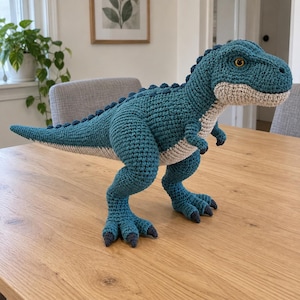 May include: A teal and cream-coloured crocheted dinosaur toy with a textured surface, a long tail, small arms, and prominent dark claws. The creature features a row of small spikes along its back and bright orange eyes, crafted with visible yarn stitches.