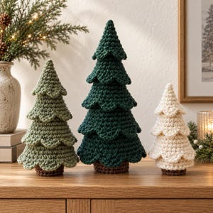 Könnte beinhalten: Drei dekorative gehäkelte Weihnachtsbäume in verschiedenen Größen und Farben. Der linke Baum ist salbeigrün, der mittlere Baum ist dunkelwaldgrün und der rechte Baum ist cremeweiß. Jeder Baum hat ein gestuftes, gewelltes Design und einen braunen Fuß.