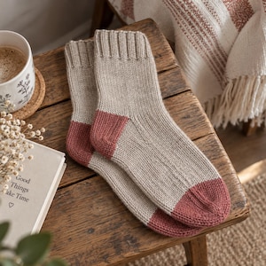 May include: A pair of knitted socks featuring a light beige main body with contrasting dark rose-coloured heels and toes. The socks have a ribbed cuff at the top and a smooth knit texture throughout the foot and ankle area, showcasing a simple design.