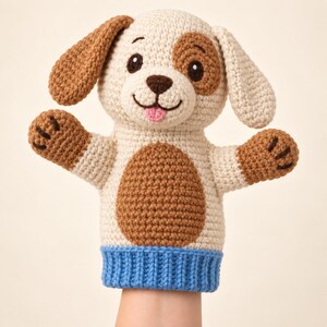 Könnte beinhalten: Eine gehäkelte Handpuppe in Form eines Hundes aus cremefarbenem Garn mit braunen Ohrflecken, einem braunen Augenfleck und einem braunen Bauchfleck. Der Hund hat schwarze gestickte Augen, eine dunkelbraune Nase, eine rosa Zunge und eine blaue gerippte Basis.