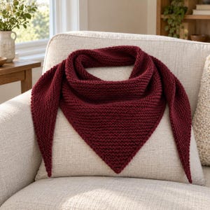 May include: A deep burgundy triangular scarf featuring a textured garter stitch knit pattern. The scarf has a wide center section that tapers into two long, pointed ends, creating a classic shawl shape with a consistent dark red yarn color.
