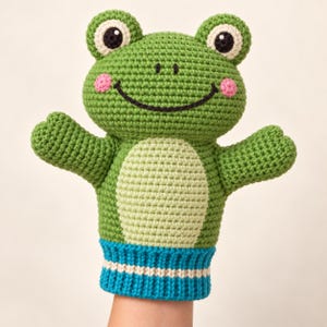 Könnte beinhalten: Eine grüne Häkel-Handpuppe in Froschform mit hellgrünem Bauch, rosa Wangen und großen Augen mit schwarzen Pupillen. Die Puppe hat ein breites, schwarz gesticktes Lächeln und ein blaues, geripptes Bündchen am unteren Rand mit einem weißen Streifen.