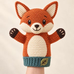 May include: A crochet fox hand puppet featuring a vibrant orange head and body with a cream-coloured muzzle and belly. The puppet has dark brown paws with light stitching, pointed ears, and a teal base adorned with a small, embroidered green leaf.