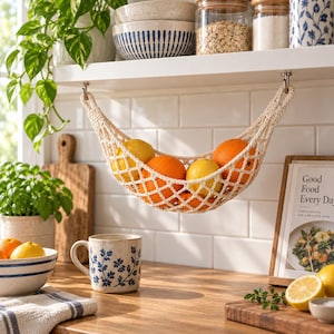 Puede incluir: Una hamaca para frutas de macramé en color crema suspendida de un estante blanco, que contiene varias naranjas y limones frescos. La red tejida presenta un patrón de diamantes y está sujeta al estante con dos pequeños ganchos metálicos.