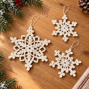 May include: Three off-white crochet snowflake ornaments with intricate patterns and loops for hanging. Each snowflake features a six-pointed star design with delicate stitching, crafted from yarn, and arranged on a wooden surface.