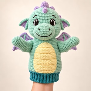 Pode incluir: Uma marioneta de mão em croché com formato de dragão, corpo verde-menta, barriga amarelo-pálido e detalhes roxos nas asas, orelhas e cristas. A marioneta apresenta olhos pretos bordados e uma boca sorridente numa cabeça arredondada.