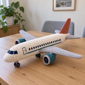 Puede incluir: Un juguete de avión tejido a ganchillo a mano con un fuselaje color crema, alas gris claro y una aleta de cola color óxido. El diseño incluye ventanas de cabina azul oscuro, una fila de pequeñas ventanas circulares y motores color verde azulado.