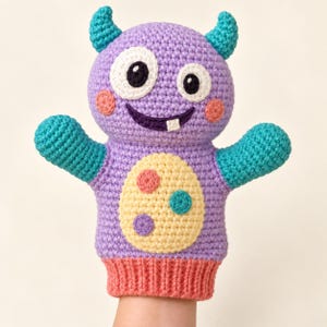 Könnte beinhalten: Eine gehäkelte Handpuppe in Form eines lila Monsters mit türkisfarbenen Hörnern und Armen. Sie hat zwei weiße Augen mit schwarzen Pupillen, einen dunkelvioletten Mund mit einem weißen Zahn, rosa Wangen und einen cremefarbenen Bauch mit Punkten.