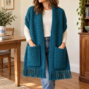 May include: A teal blue crochet vest featuring a textured stitch pattern, two large front patch pockets, and a fringe trim along the bottom hem. The garment is worn over a long-sleeved white shirt and blue denim jeans, creating a layered look.