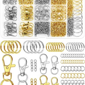 Può includere: Un kit completo per la creazione di gioielli e portachiavi con una scatola organizer in plastica trasparente riempita di moschettoni, anellini di giunzione e anelli portachiavi color argento e oro, con componenti metallici sparsi sotto.