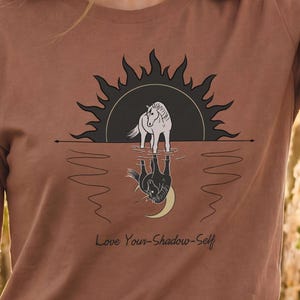 May include: A brown t-shirt featuring a graphic print of a white horse standing in water with a sun behind it and a reflection of the horse with a crescent moon. The text "Love Your-Shadow-Self" is printed in black below the graphic design.