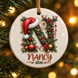 Op de afbeelding: Een ronde witte keramische kerstornament met een grote letter N versierd met dennentakken, dennenappels, rode bessen en gouden ballen. Een rode kerstmuts zit bovenop de letter. De naam NANCY en het jaartal 2026 staan eronder gedrukt.