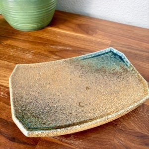 May include: A rectangular ceramic tray with a crackle glaze finish. The surface features a gradient of tan and beige tones transitioning into a deep teal blue at one end, with dark speckles throughout the textured, crazed surface pattern.