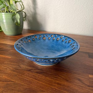 May include: A round ceramic bowl with a deep blue glaze and a series of small, circular cutouts along the flared rim. The interior of the bowl is smooth and glossy, while the exterior edge features a dark, textured finish around each opening.