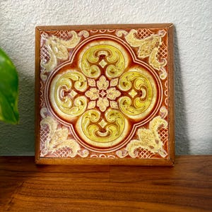 May include: A square ceramic tile featuring a raised relief pattern in shades of yellow, gold, and deep brown. The design consists of a central floral motif surrounded by four symmetrical scrolls, all encased in a simple wooden frame.