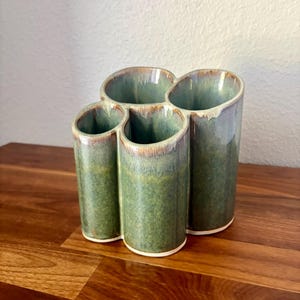 May include: A ceramic utensil holder featuring four interconnected cylindrical compartments with a mottled green glaze and brown rim accents. The structure has a smooth, glossy finish and a white base, providing a multi-sectioned storage vessel.