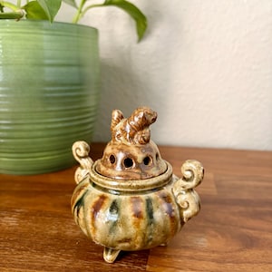 May include: A small, round, glazed ceramic incense burner featuring a brown and green mottled finish. The lid is topped with a sculpted animal figure and includes circular ventilation holes. It has two curved side handles and three small feet.