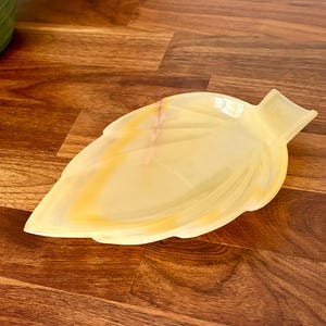 May include: A leaf-shaped dish carved from translucent yellow and cream-colored stone. The decorative tray features smooth, curved edges and subtle natural veining throughout the material, with a small stem-like handle at one end.