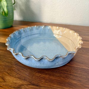 Può includere: Una tortiera rotonda in ceramica con bordo smerlato e ondulato. L'interno presenta uno smalto bicolore, con una metà in azzurro chiaro maculato e l'altra metà in beige crema liscio. Il bordo esterno mostra una sottile finitura marrone.