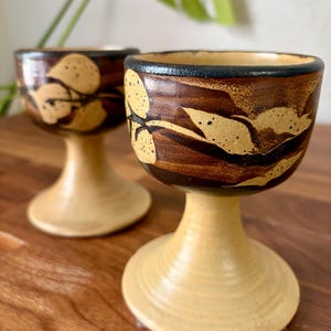 May include: Two ceramic goblets featuring a cream-colored base and stem, with a dark brown upper bowl decorated with a speckled tan leaf pattern and a black rim. The vessels have a wide, circular foot and a smooth, rounded drinking edge.