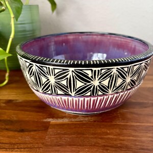 May include: A ceramic bowl featuring a deep purple interior and a black exterior band decorated with a white geometric flower pattern. The lower portion of the bowl has vertical carved ridges, and the rim is finished with a black edge.