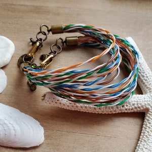 May include: A multi-strand bracelet made from twisted colourful wires with gold-toned clasps and accents. The bracelet is displayed on a white starfish and seashells.