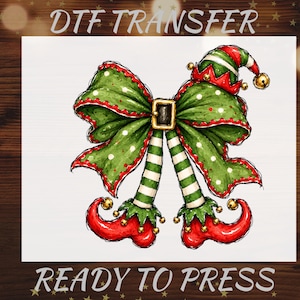 May include: A DTF TRANSFER READY TO PRESS graphic featuring a large green bow with white polka dots and red trim, centered with a black belt buckle. Below the bow are two striped green and white legs ending in curled red elf shoes with gold bells.